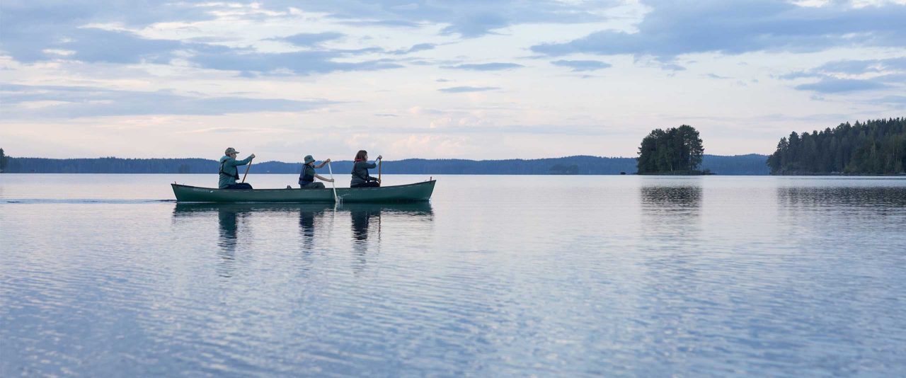 Kolme melojaa meloo kanootissa. Järvi on tyyni ja taivaalla näkyy auringonlaskun sävyjä.