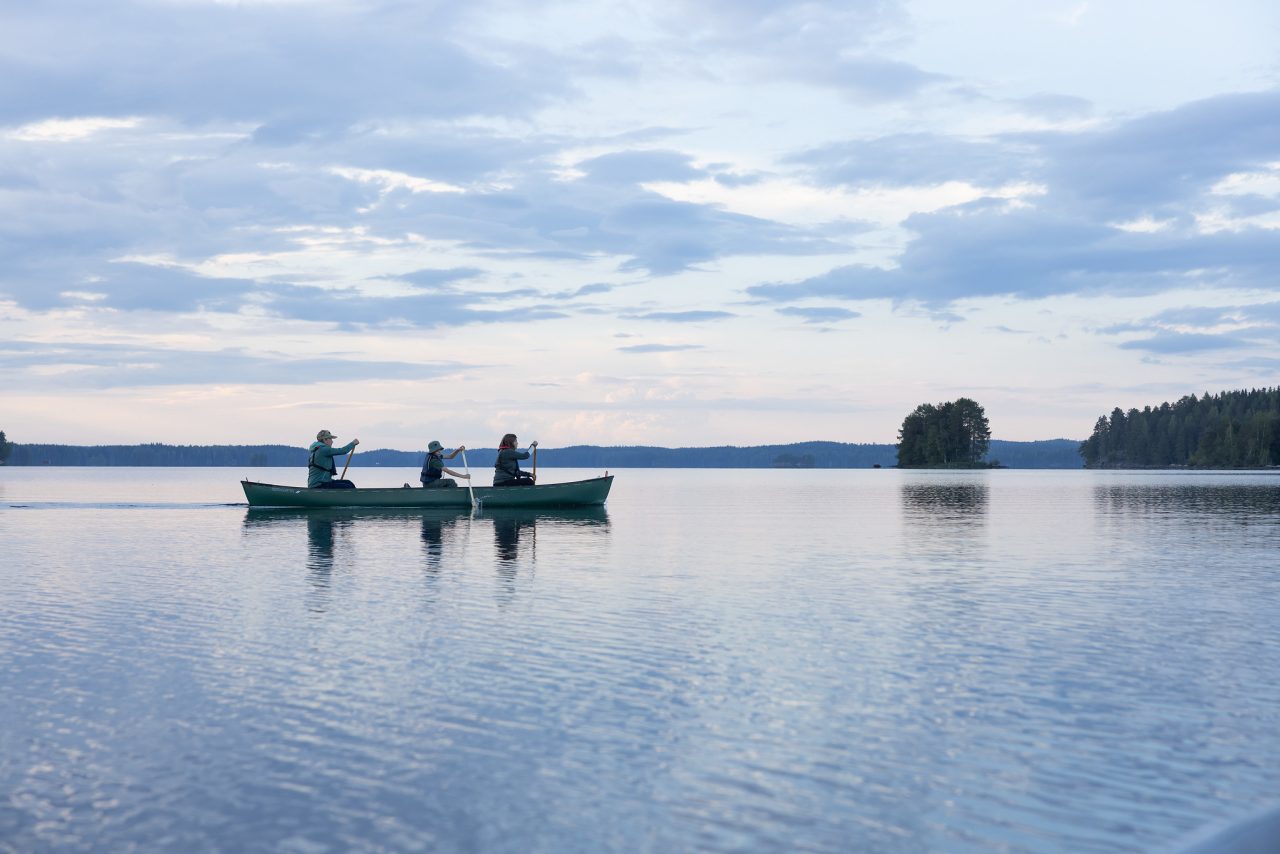 Kolme ihmistä kanootissa tyynellä järvellä auringonlaskun aikaan.