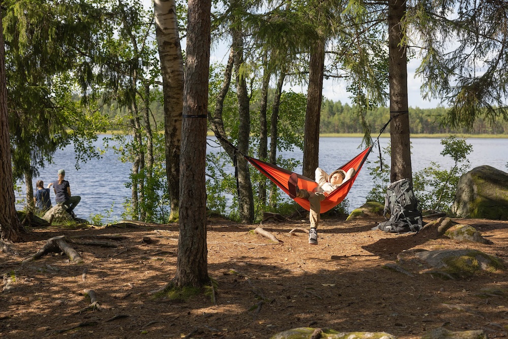 Mies rentoutuu riippumatossa järvimaisemassa metsässä.