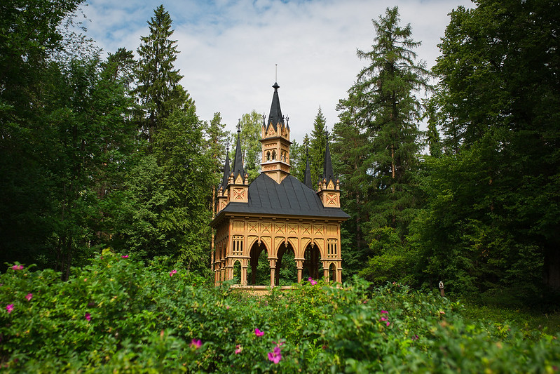 Keltainen koristeellinen puusta rakennettu paviljonki metsän keskellä. Edustalla on tumman vaaleanpunaisia ruusuja.