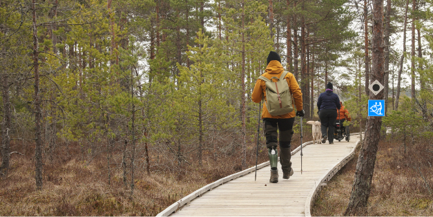 Sauvakävelijä, pyörätuolilla liikkuva ja avustajakoiran kanssa liikkuva ihminen leveällä pitkospuupolulla.